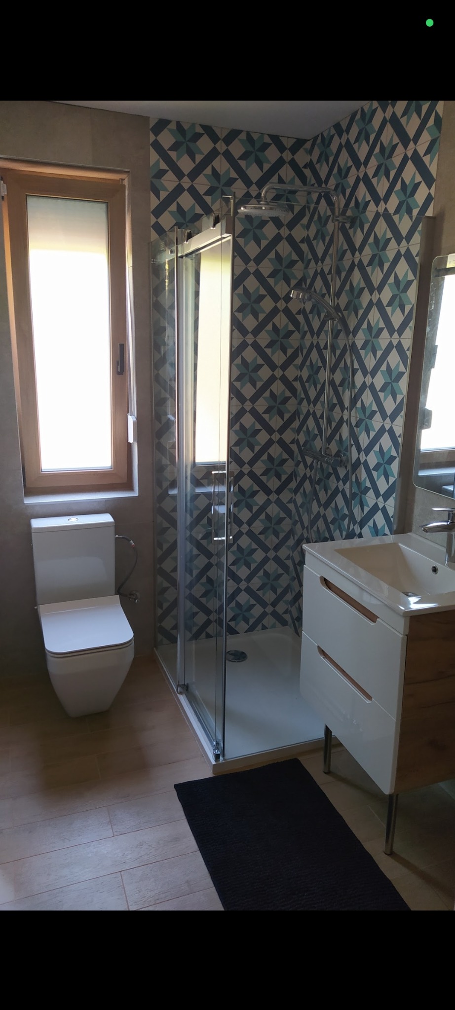 Wykończona łazienka z kabiną prysznicową, toaletą i umywalką z szafką. Ściany wyłożone płytkami w geometryczne wzory. Na podłodze drewnopodobne płytki i czarny dywanik.