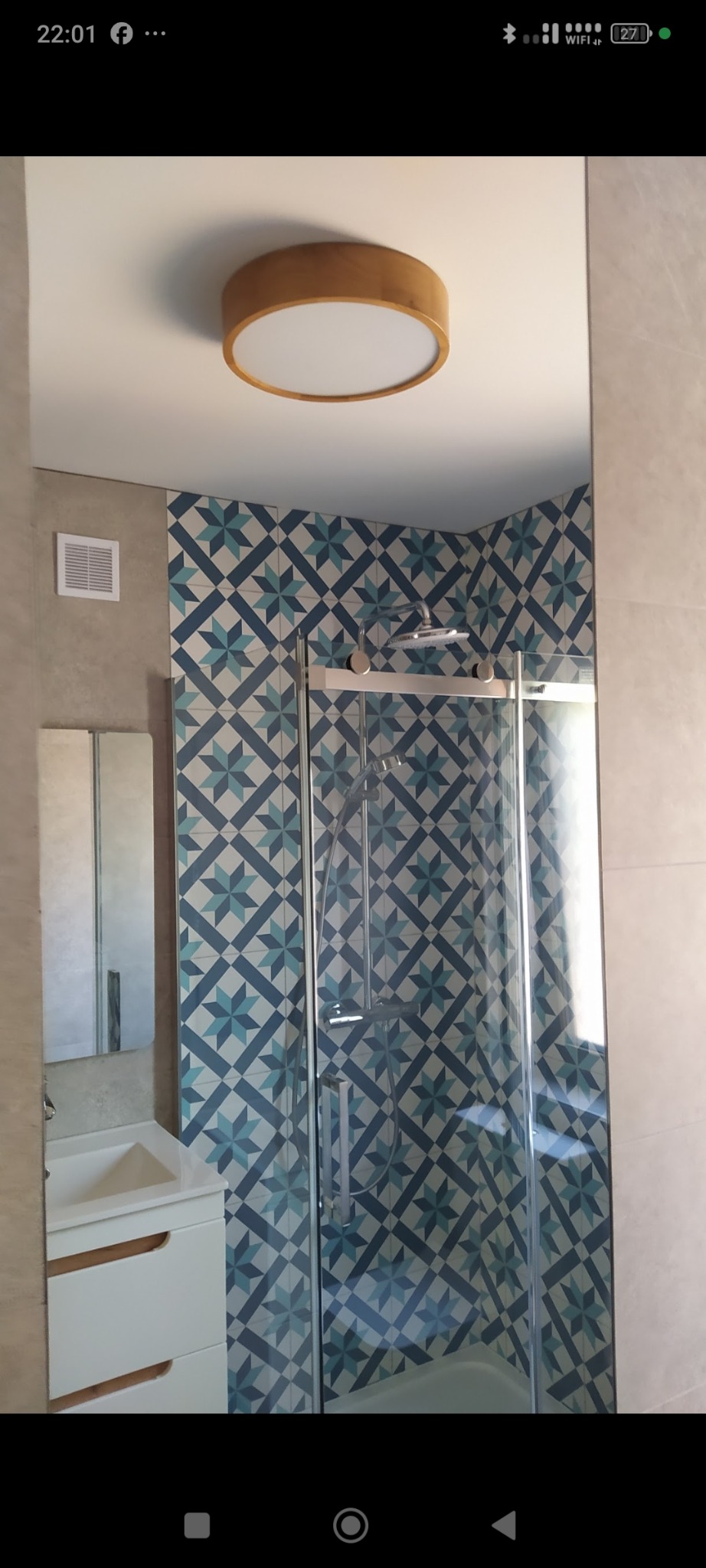 Wykończona łazienka z kabiną prysznicową za szklanymi drzwiami, wzorzyste płytki w odcieniach błękitu i beżu, okrągła lampa sufitowa z drewnianą obudową i biała szafka z umywalką.