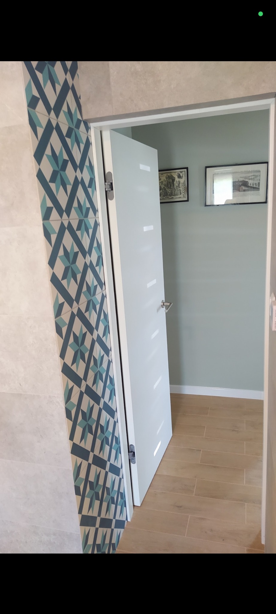 Wykończona łazienka z białą, nowoczesną stolarką drzwiową i dekoracyjnymi płytkami w geometryczne wzory. Widoczna podłoga z płytek imitujących drewno i ściany w odcieniach szarości.