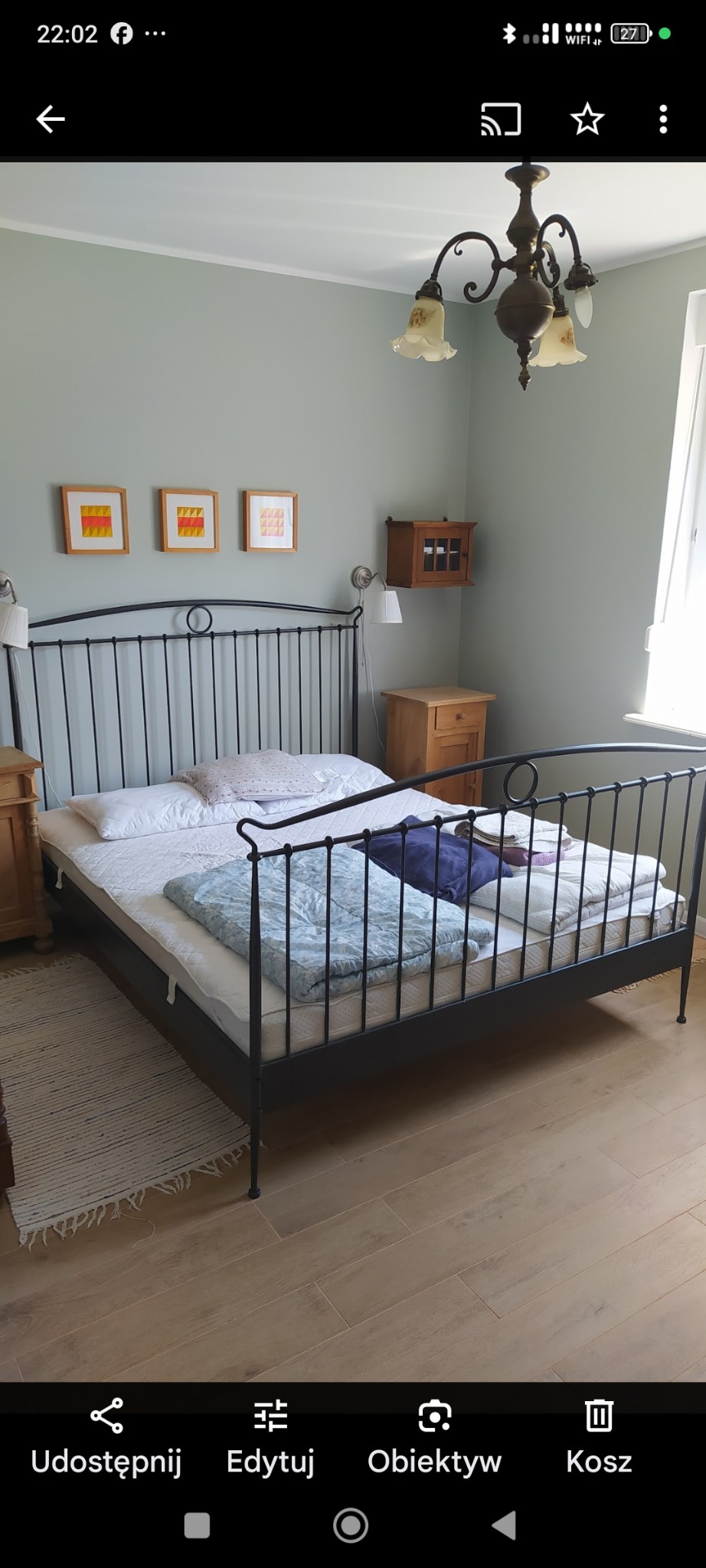 Jasna sypialnia z metalowym łóżkiem, drewnianą podłogą i trzema obrazkami na ścianie. Na ścianie wisi mała szafka. Klasyczny żyrandol.