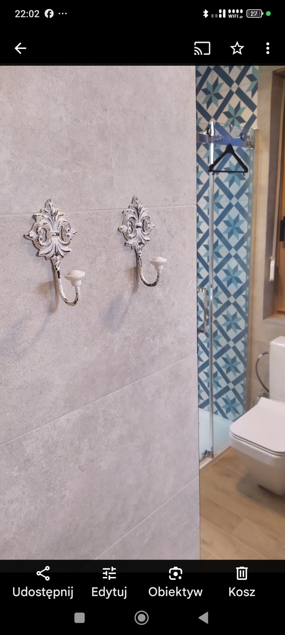 Wykończona łazienka z szarymi płytkami, dekoracyjnymi wieszakami w stylu vintage i kabiną prysznicową z niebieską mozaiką. Widoczny fragment toalety i drewnopodobna podłoga.