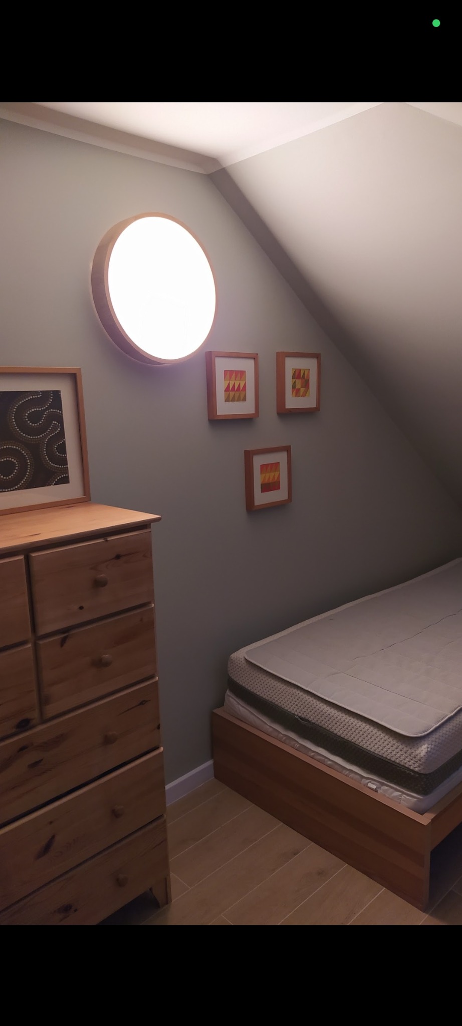 Pokój na poddaszu z drewnianą komodą, łóżkiem i okrągłą lampą na ścianie. Na ścianie wiszą trzy małe obrazki w drewnianych ramkach. Ukośny sufit.