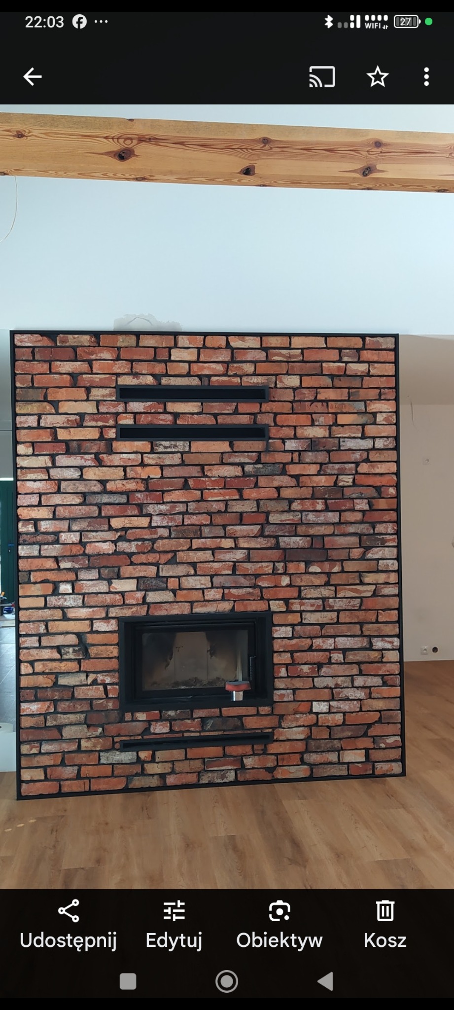 Wykończony kominek obłożony cegłą dekoracyjną z czarną ramą i wkładem kominkowym. Widoczne drewniane belki stropowe i podłoga z paneli.