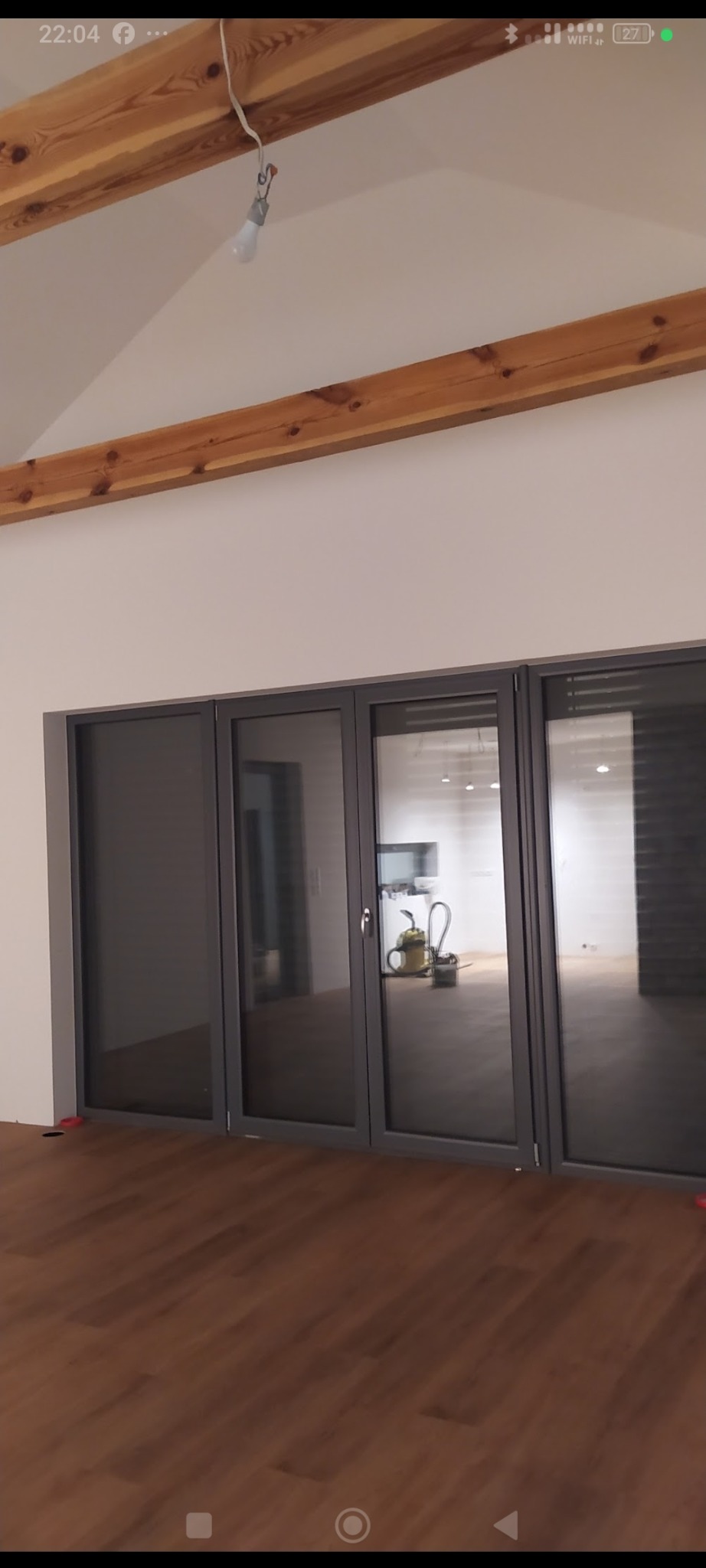 Nowoczesne, czteroskrzydłowe drzwi balkonowe z szarymi ramami, widok na wnętrze z drewnianą podłogą i belkami stropowymi. W tle widoczny odkurzacz.