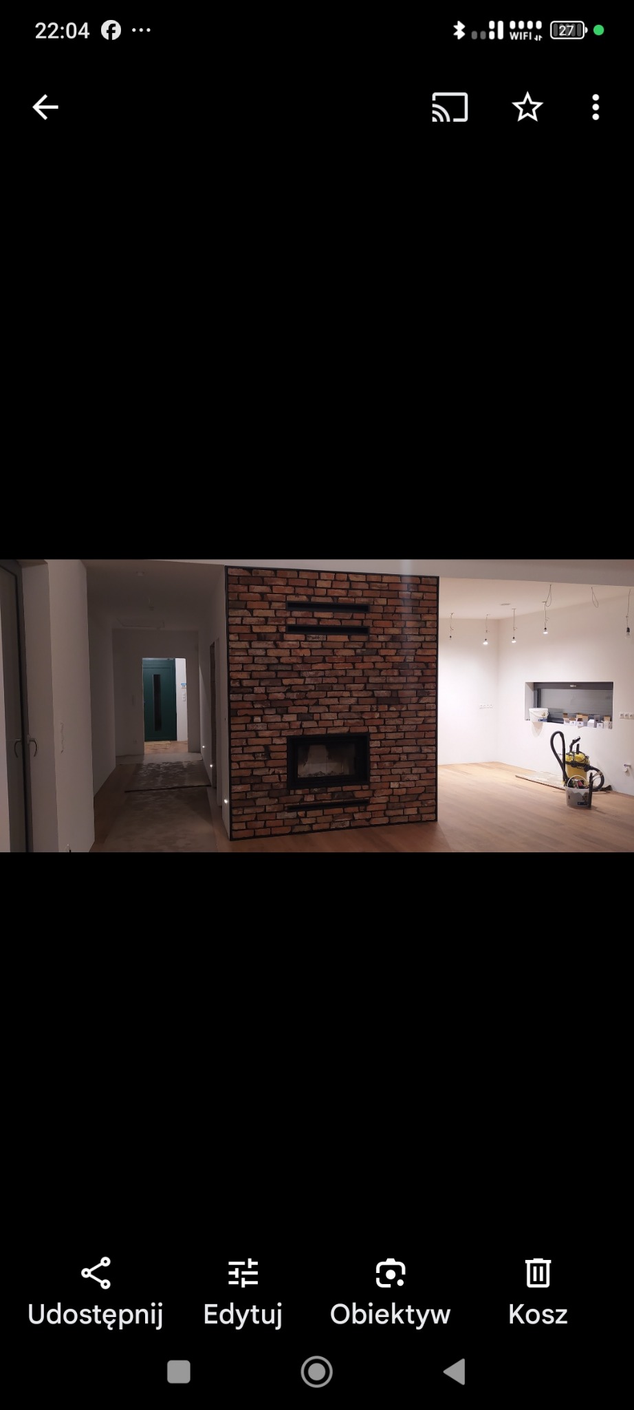 Salon po remoncie z kominkiem obłożonym cegłą, parkiet na podłodze, widoczne ślady po remoncie, odkurzacz i wiadro z farbą. Minimalistyczny design.