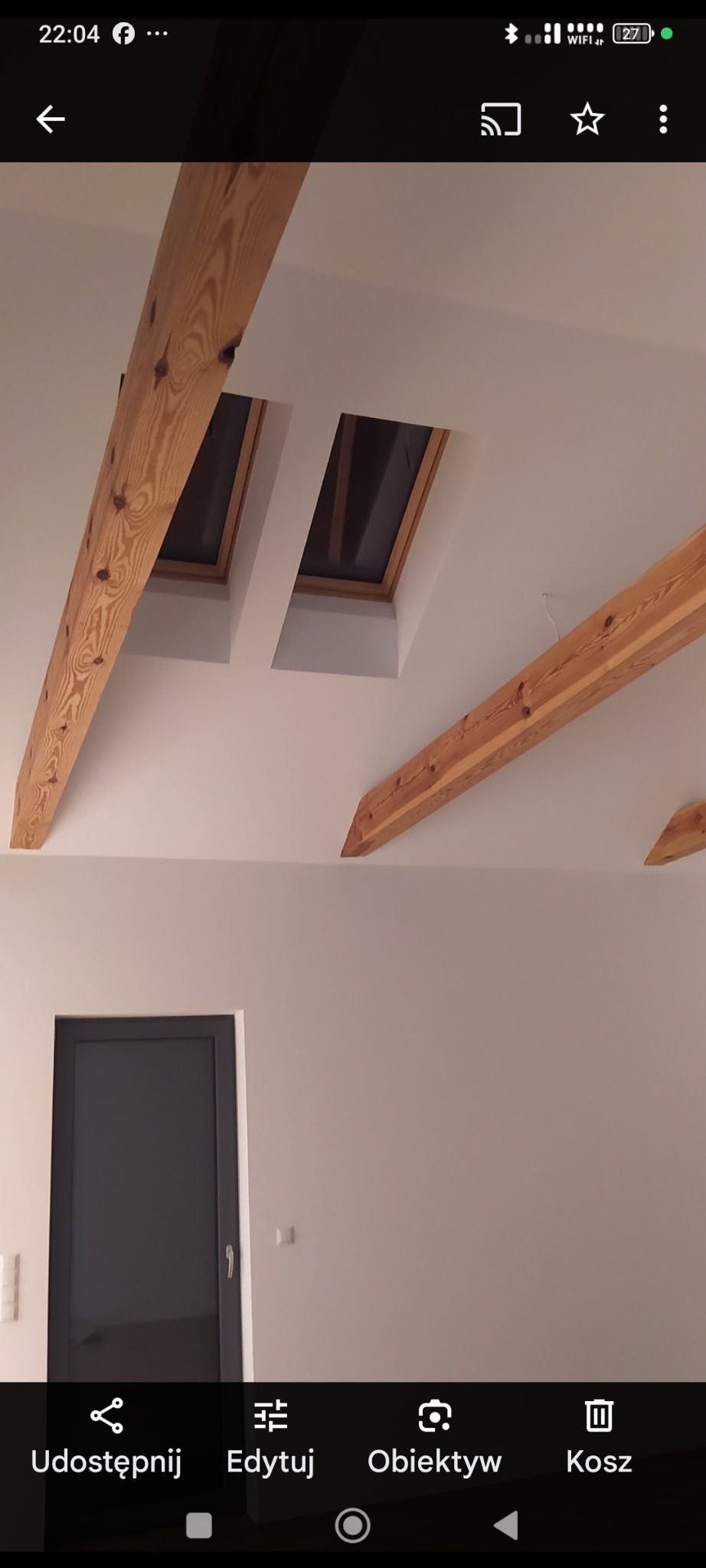 Jasne wnętrze poddasza z drewnianymi belkami stropowymi i dwoma oknami dachowymi. Widoczna ściana z drzwiami w ciemnej ramie. Minimalistyczny design.