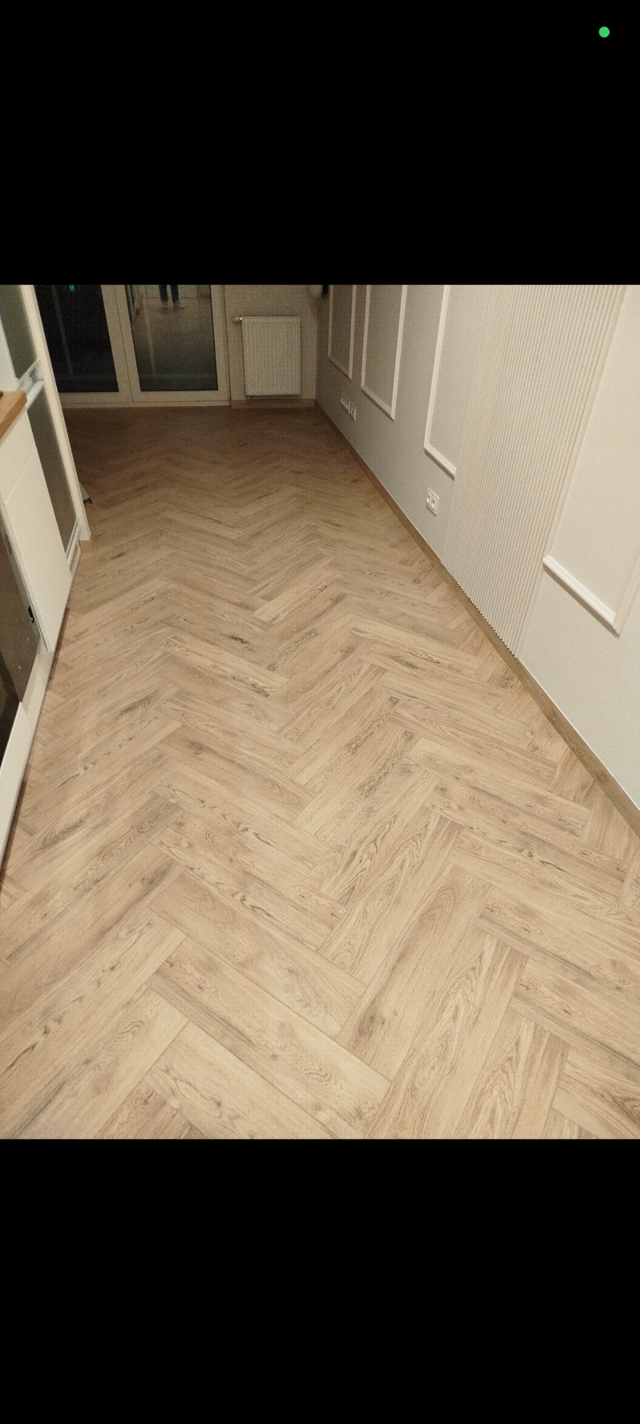 Jasna, drewniana podłoga ułożona w jodełkę francuską, świeżo po montażu. Widoczne listwy przypodłogowe i fragment ściany z dekoracyjnymi panelami.