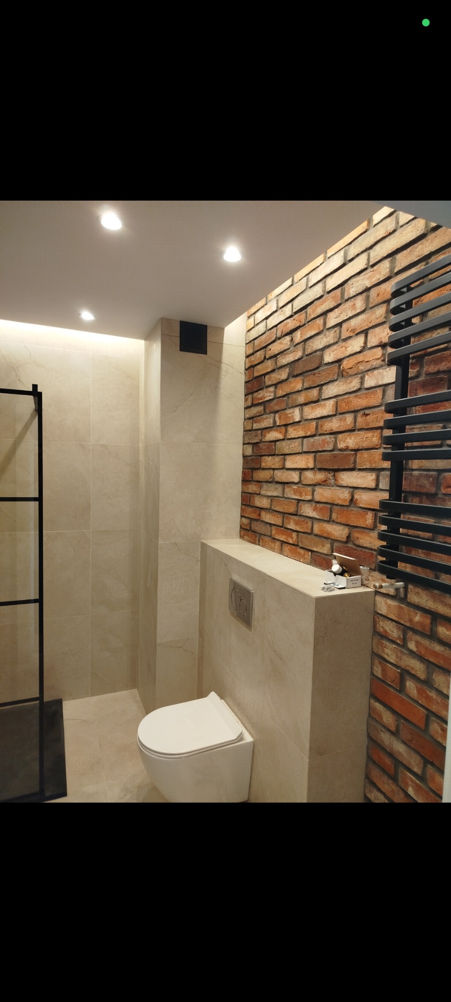 Nowoczesna łazienka z toaletą podwieszaną, beżowymi płytkami i ścianą z czerwonej cegły. Grzejnik drabinkowy w kolorze czarnym. Oświetlenie punktowe.