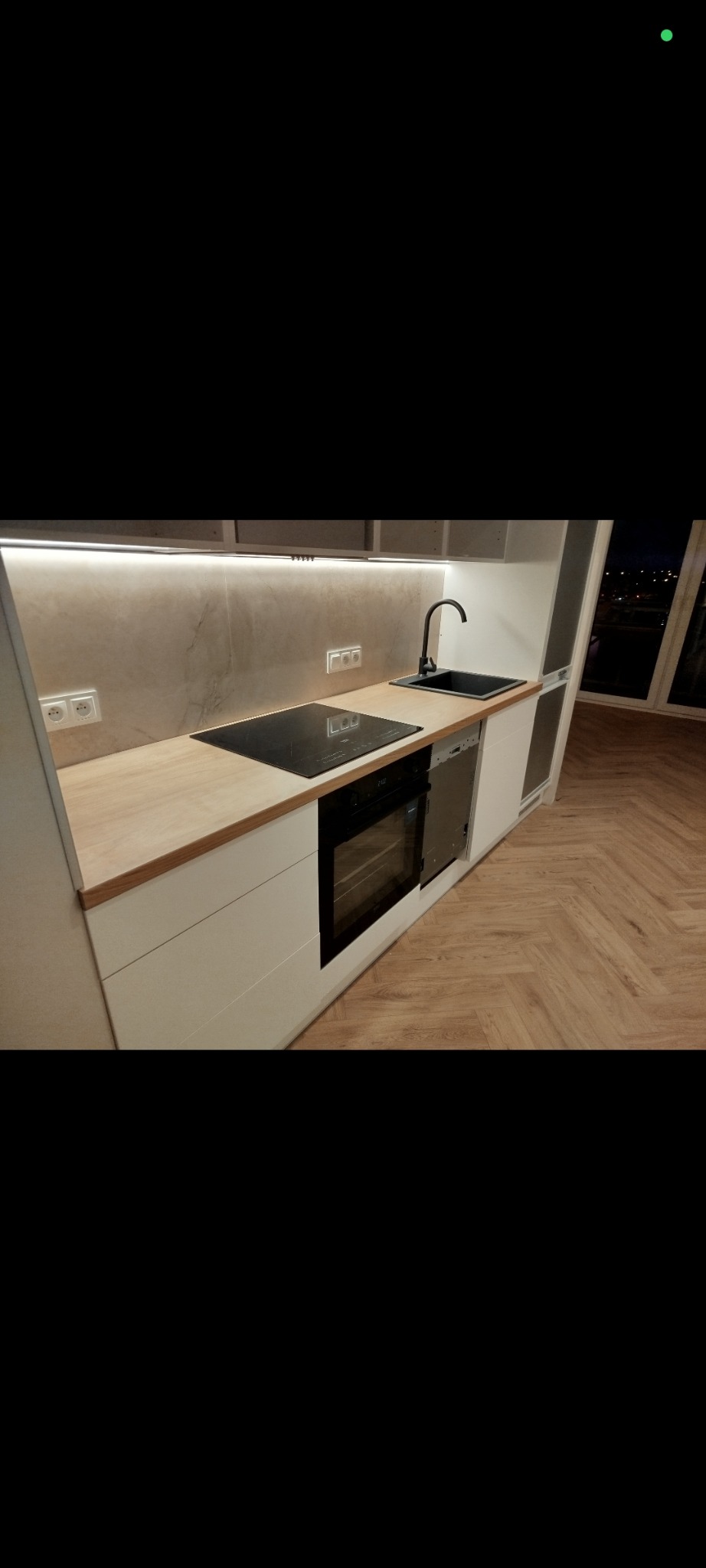 Nowoczesna kuchnia z białymi szafkami, drewnianym blatem i czarną armaturą. Uwagę przykuwa parkiet ułożony w jodełkę francuską i subtelne oświetlenie LED pod szafkami.