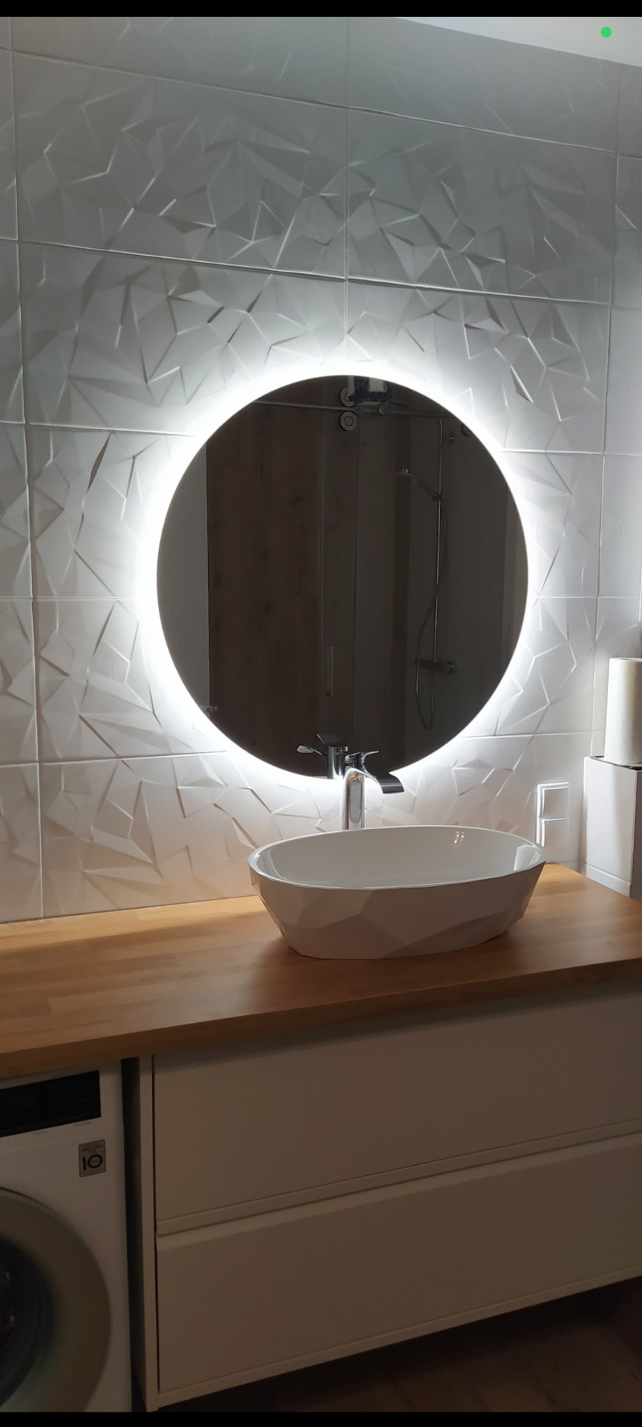 Nowoczesna łazienka z umywalką nablatową, okrągłym lustrem z podświetleniem LED i trójwymiarowymi białymi płytkami na ścianie. Widoczna pralka LG.