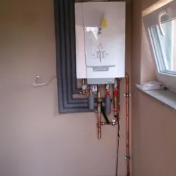 Biały piec gazowy De Dietrich zamontowany na ścianie z miedzianymi rurami, z izolacją termiczną na rurach doprowadzających, w pomieszczeniu z oknem.