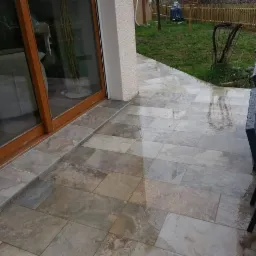 Wyłożony płytkami taras przy domu z przesuwnymi drzwiami balkonowymi, widoczne krople deszczu na powierzchni płytek i meblach ogrodowych.