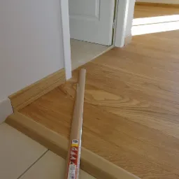 Połączenie podłogi drewnianej z płytkami ceramicznymi, widoczne listwy przypodłogowe z drewna, jedna jeszcze w folii ochronnej, leżąca obok miejsca montażu.