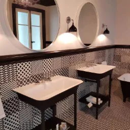 Nowoczesna łazienka w czarno-białej kolorystyce: dwie umywalki na czarnych postumentach, okrągłe i owalne lustra z czarnymi lampami, wzorzyste płytki na ścianach i podłodze, widoczna wanna.