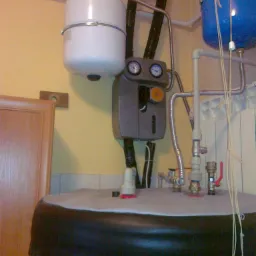 Instalacja centralnego ogrzewania z dwoma zbiornikami ciśnieniowymi (białym i niebieskim), manometrami, zaworami i rurami w pomieszczeniu technicznym o żółtych ścianach i częściowo wykończonymi...