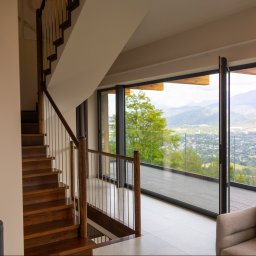 PPUH Art-Rad - Nowoczesne wnętrze z drewnianymi schodami i panoramicznym oknem z widokiem na góry. Minimalistyczny design, jasne kolory, kominek.