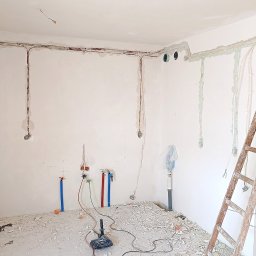 PPUH Art-Rad - Remont pokoju w trakcie: instalacja elektryczna i hydrauliczna w bruzdach, drabina malarska, resztki gruzu na podłodze. Widoczne kable i rury.