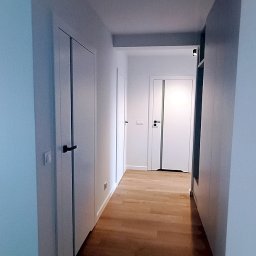 PPUH Art-Rad - Nowoczesny korytarz z drewnianą podłogą i białymi drzwiami z czarnymi klamkami. Minimalistyczny design, jasne ściany, perspektywa w głąb mieszkania.