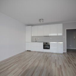 PPUH Art-Rad - Nowoczesna, minimalistyczna kuchnia z białymi, lakierowanymi frontami szafek i drewnianą podłogą. Otwarta przestrzeń, widok na sąsiednie pomieszczenie przez otwarte drzwi.