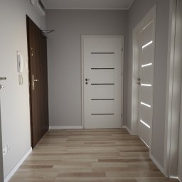 PPUH Art-Rad - Jasne wnętrze przedpokoju z trzema parami drzwi: ciemne wejściowe, białe z poziomymi frezami i białe z pionowymi. Podłoga wyłożona panelami imitującymi drewno.