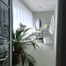 PPUH Art-Rad - Jasna łazienka z wanną, umywalką na metalowym stelażu, toaletą i rośliną w koszu. Minimalistyczny design z szarymi płytkami i białymi zasłonami.