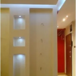 Ściana z wnękami podświetlonymi punktowym oświetleniem LED, widoczne ślady po instalacji elektrycznej, fragment korytarza z drzwiami i czerwonym akcentem kolorystycznym w tle.