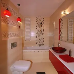 Jasna łazienka z beżowymi płytkami, dekoracyjnym pasem z motywem kwiatowym, podwieszaną toaletą, czerwonymi lampami wiszącymi, umywalką w kształcie misy i szafką w kolorze czerwieni.