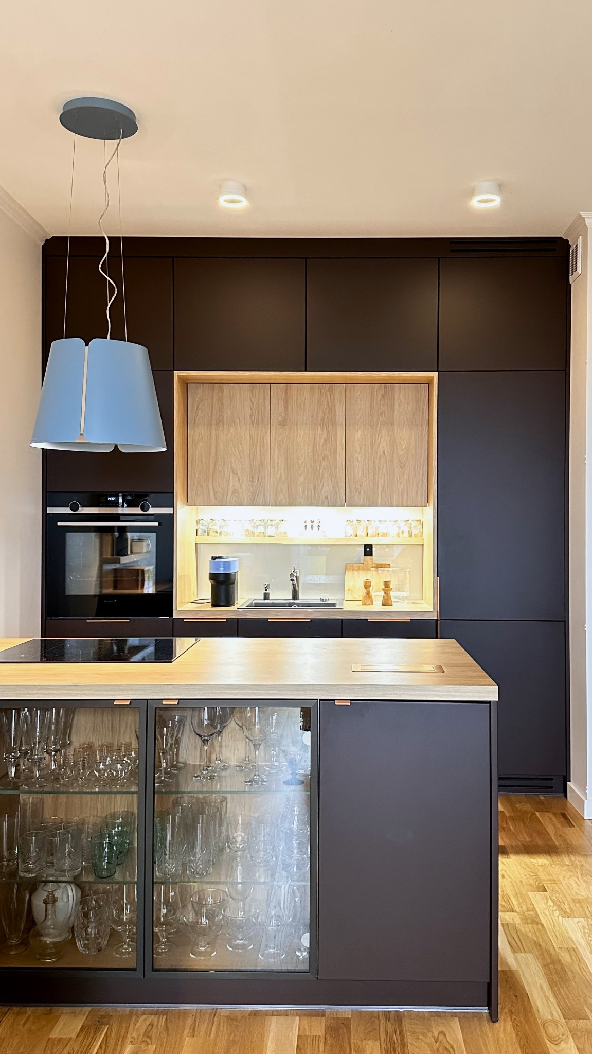 Nowoczesna kuchnia z ciemnymi szafkami, drewnianym blatem i wyspą z przeszklonymi drzwiami. Oświetlenie LED podkreśla detale i dodaje elegancji.