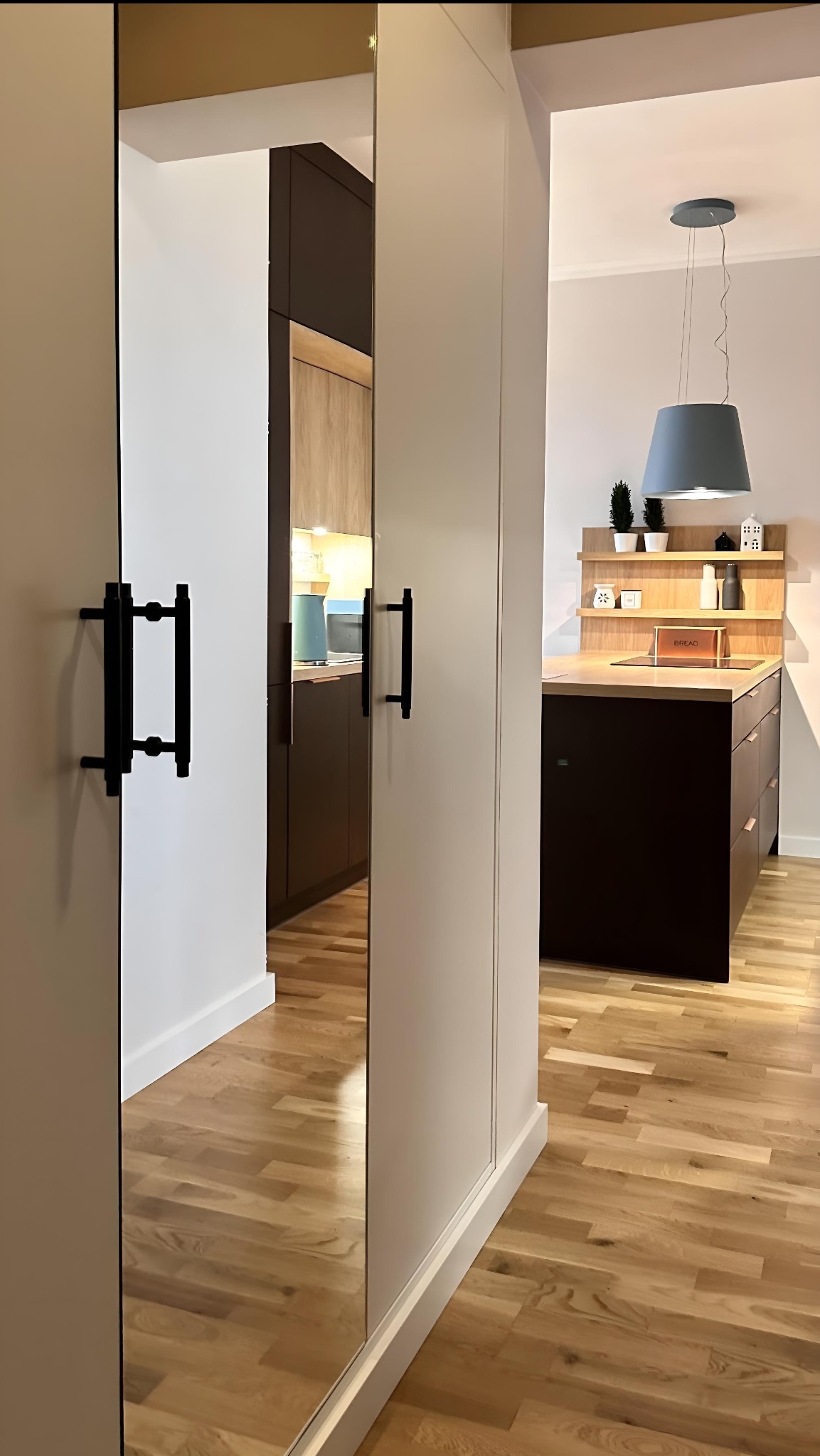 Nowoczesna kuchnia z wyspą i lustrzanymi drzwiami szafy w przedpokoju. Widoczne drewniane podłogi i minimalistyczne oświetlenie.