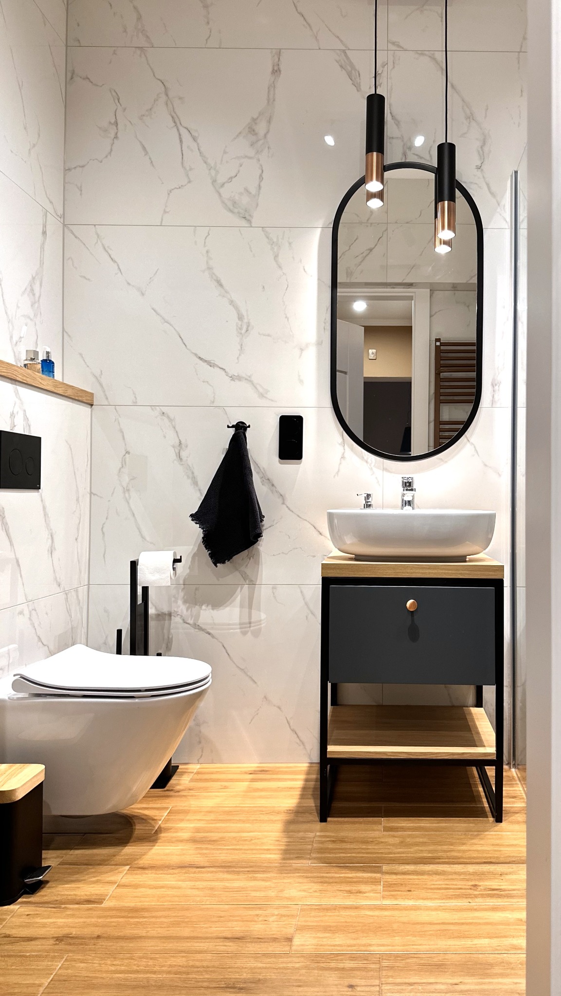 Nowoczesna łazienka z marmurowymi płytkami, drewnianą podłogą i owalnym lustrem. Uwagę przykuwają minimalistyczne lampy wiszące i designerska umywalka.