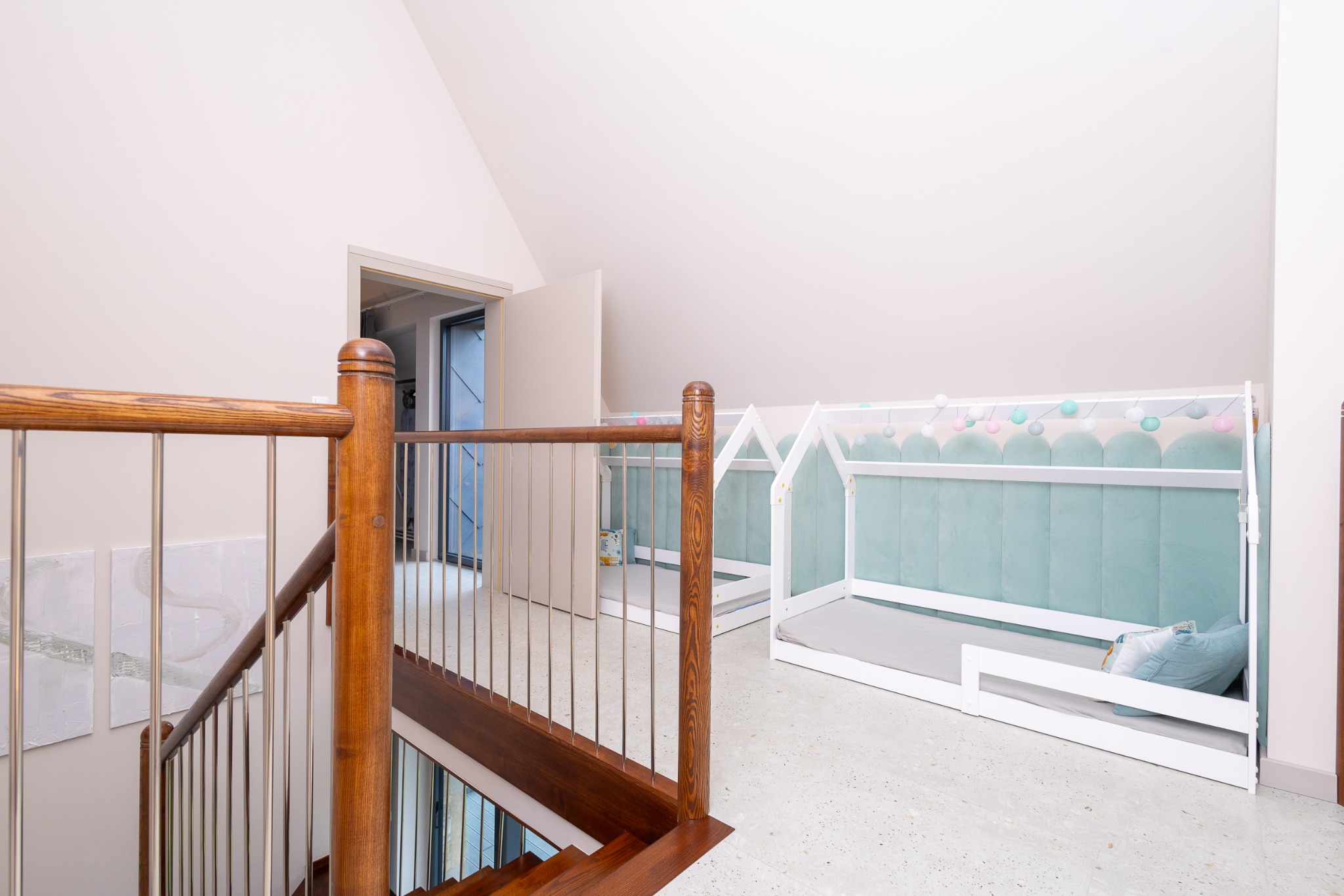 Jasne poddasze z drewnianymi schodami i dwoma białymi łóżkami w kształcie domków, wykończone pastelową tapicerką. Widok z góry na nowoczesne wnętrze.