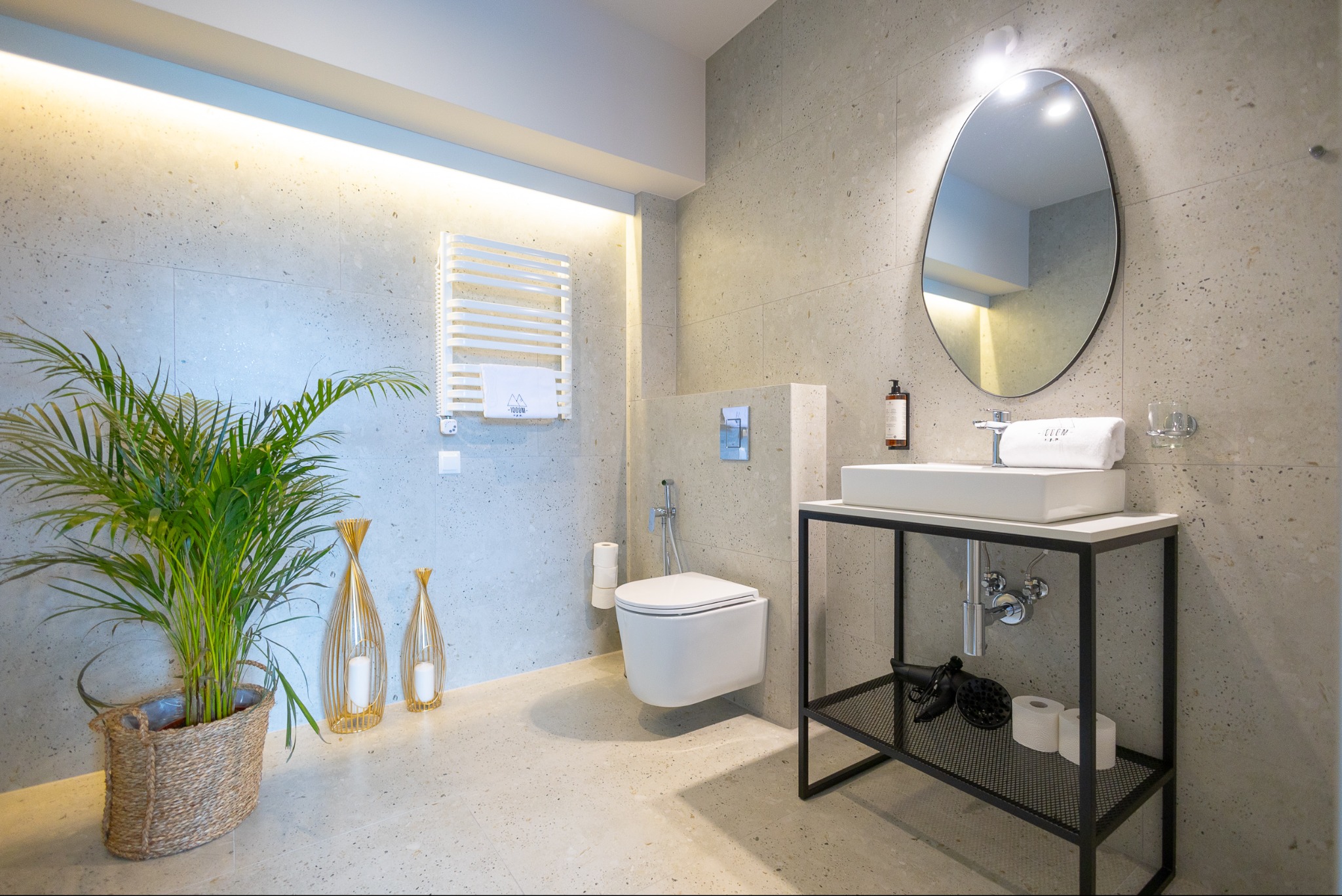 Stylowa łazienka z terazzo: wisząca toaleta, umywalka na czarnym stelażu, grzejnik drabinkowy i dekoracje. Oświetlenie LED podkreśla fakturę ścian.