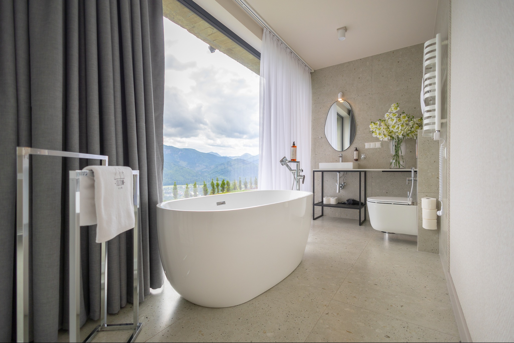 Nowoczesna łazienka z wolnostojącą wanną na tle okna z widokiem na góry. Minimalistyczny design, jasne kolory, kamienna posadzka i eleganckie dodatki.