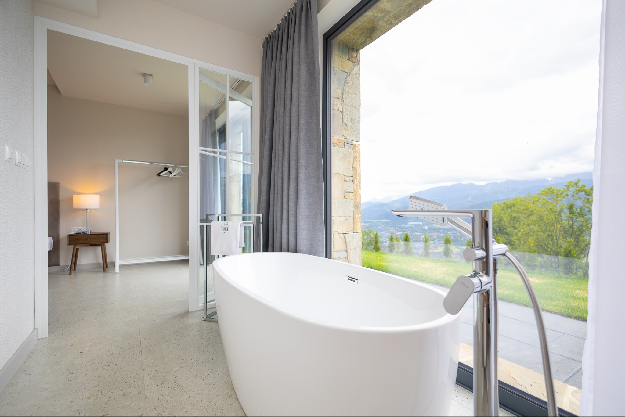 Nowoczesna, biała wanna wolnostojąca z chromowaną armaturą na tle panoramicznego okna z widokiem na góry. Minimalistyczny design łazienki z elementami kamienia i drewna.