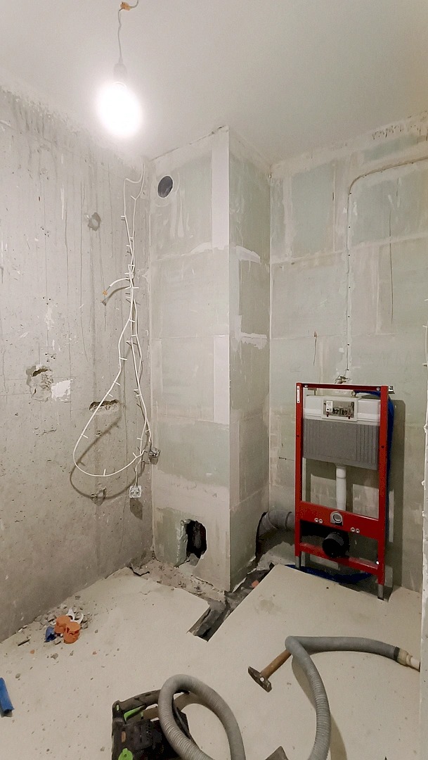 Wnętrze łazienki w trakcie remontu: surowe ściany, instalacja podtynkowa WC na czerwonym stelażu, widoczne przewody elektryczne i narzędzia. Etap przygotowania do wykończenia.