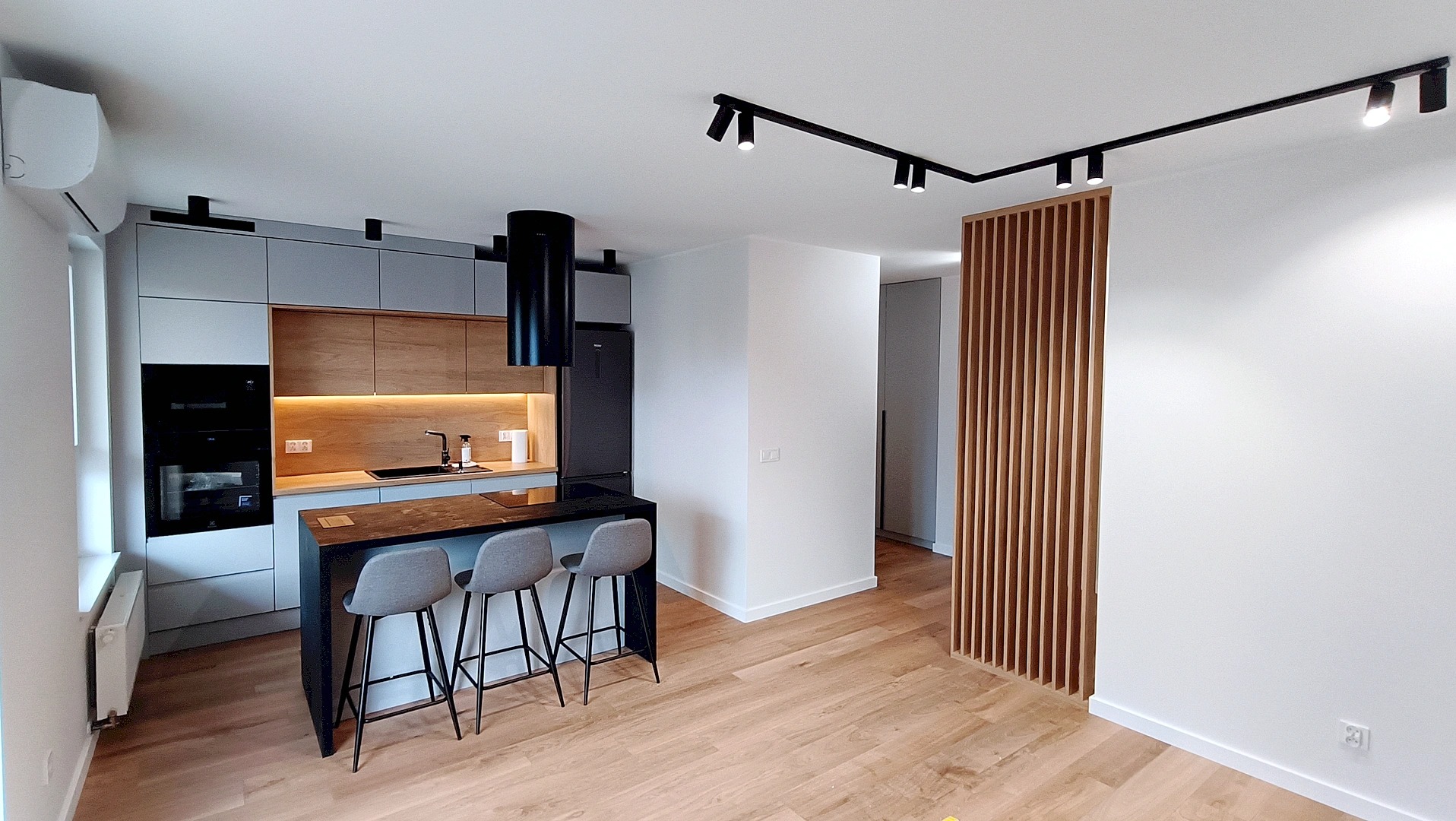 Nowoczesna kuchnia z wyspą i drewnianą ścianką działową. Szare szafki, czarny okap i oświetlenie szynowe. Jasna podłoga i minimalistyczny design.