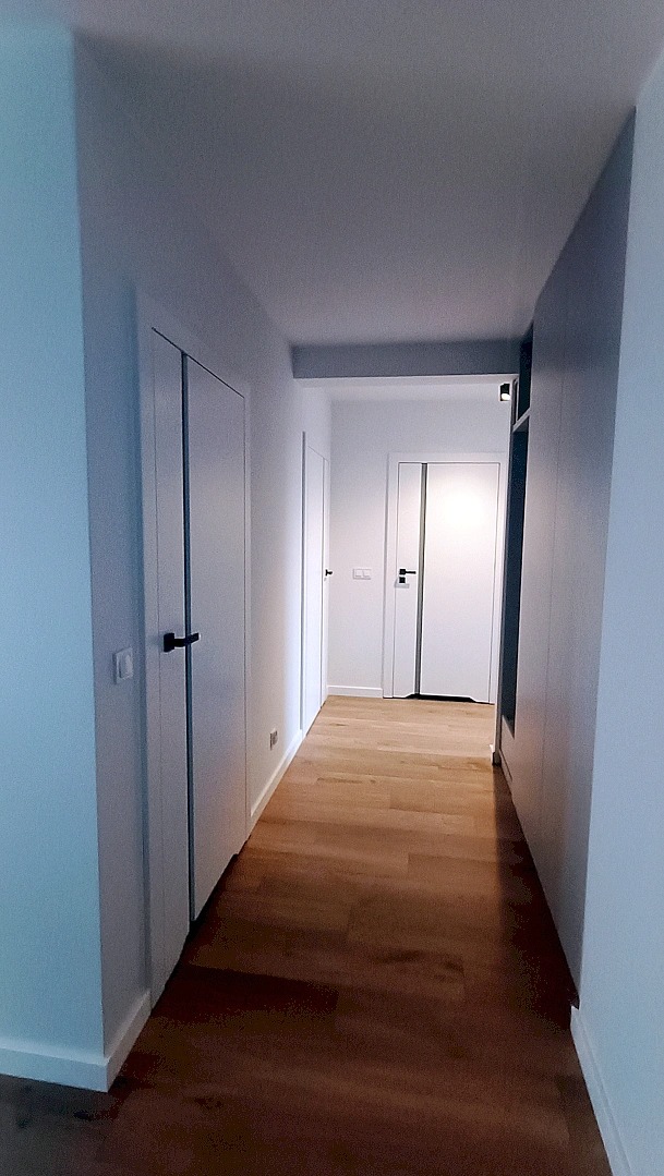 Nowoczesny korytarz z drewnianą podłogą i białymi drzwiami z czarnymi klamkami. Minimalistyczny design, jasne ściany, perspektywa w głąb mieszkania.