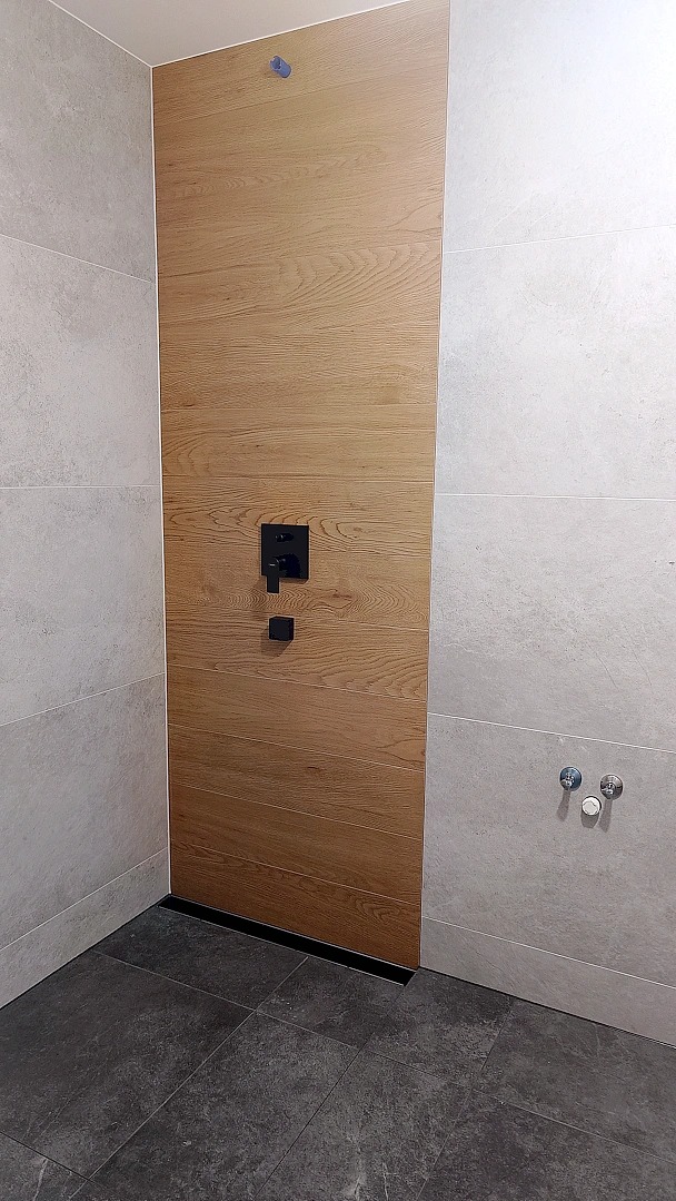 Nowoczesna kabina prysznicowa z drewnopodobną ścianą i czarną armaturą. Szare płytki na ścianach i podłodze. Minimalistyczny design, widoczne detale wykończenia.