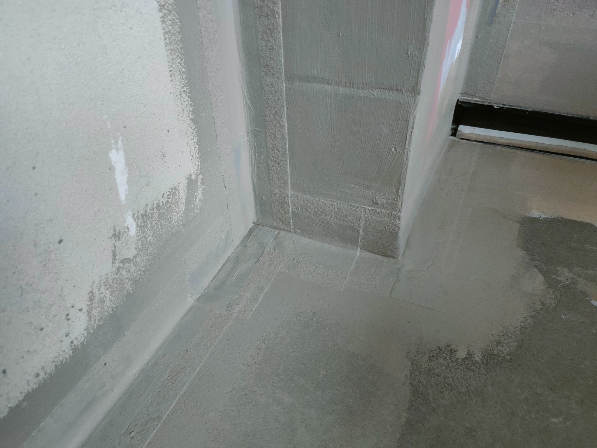 Szara, świeżo nałożona hydroizolacja w rogu łazienki, widoczne ślady pędzla i taśmy malarskiej. Przygotowanie do dalszych prac wykończeniowych.