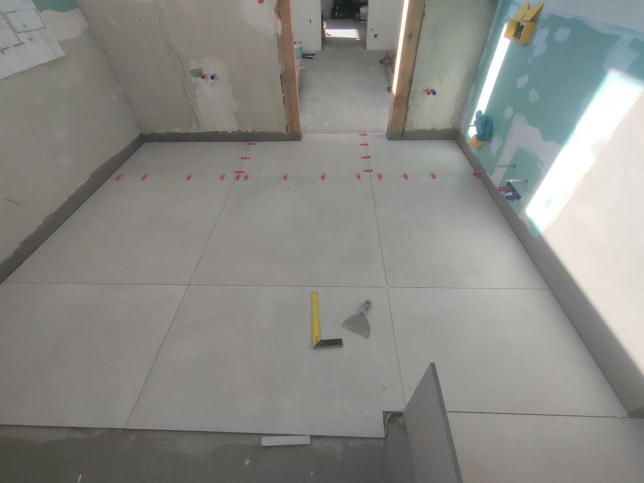 Układanie jasnych, kwadratowych płytek podłogowych w pomieszczeniu w trakcie remontu. Widoczne narzędzia: poziomica, szpachelka i elementy do poziomowania płytek.