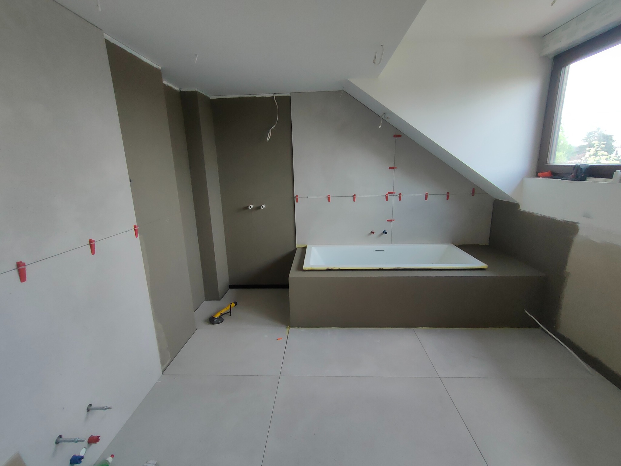 Łazienka w trakcie wykończenia: biała wanna obudowana szarą konstrukcją, ściany przygotowane do położenia płytek, widoczne narzędzia i poziomica. Podłoga wyłożona jasnymi płytkami.