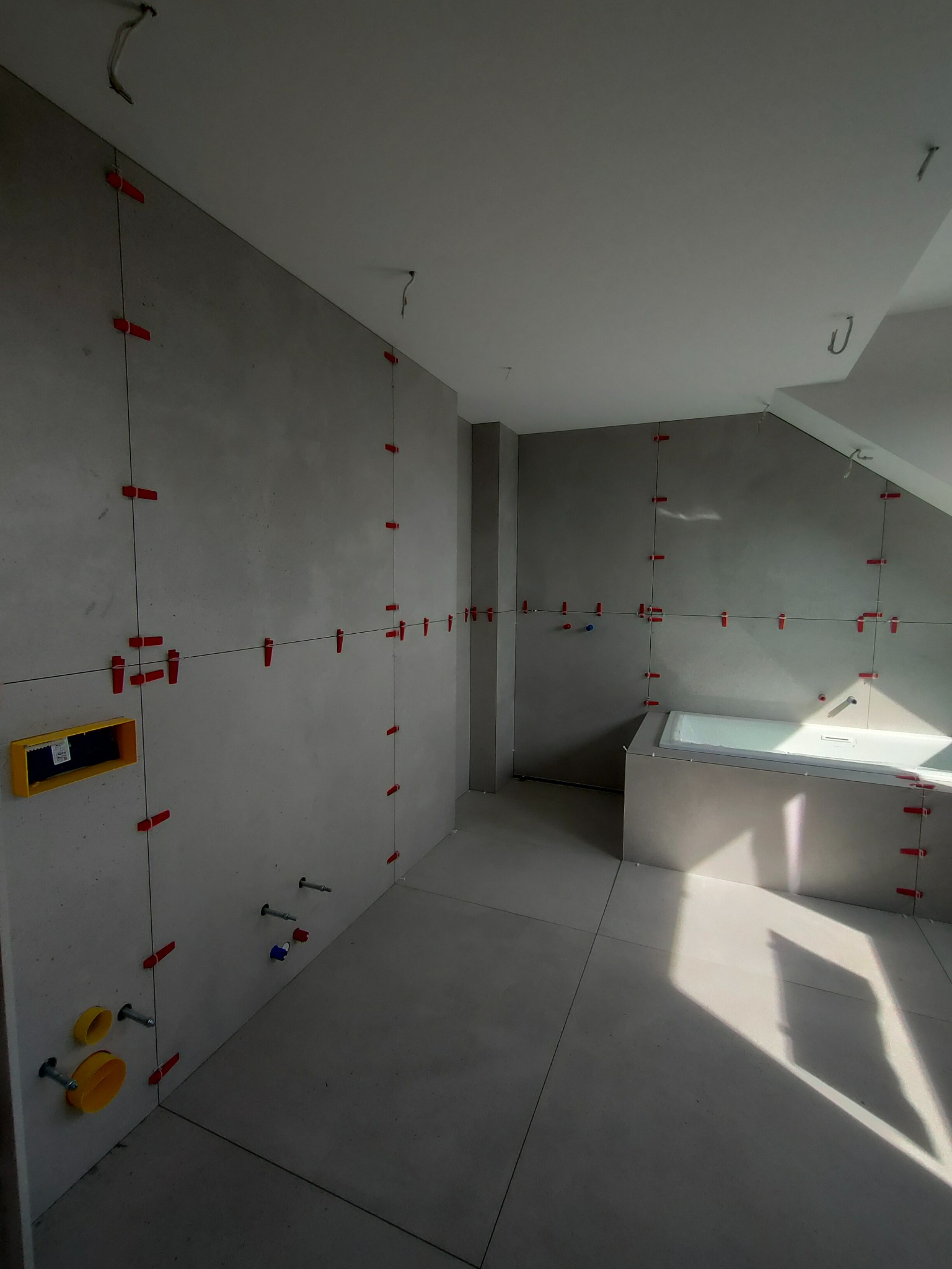Łazienka w trakcie wykańczania: szare płytki, czerwone krzyżyki dystansowe, widoczne instalacje wodne i fragment wanny. Ukośny sufit z oknem dachowym.