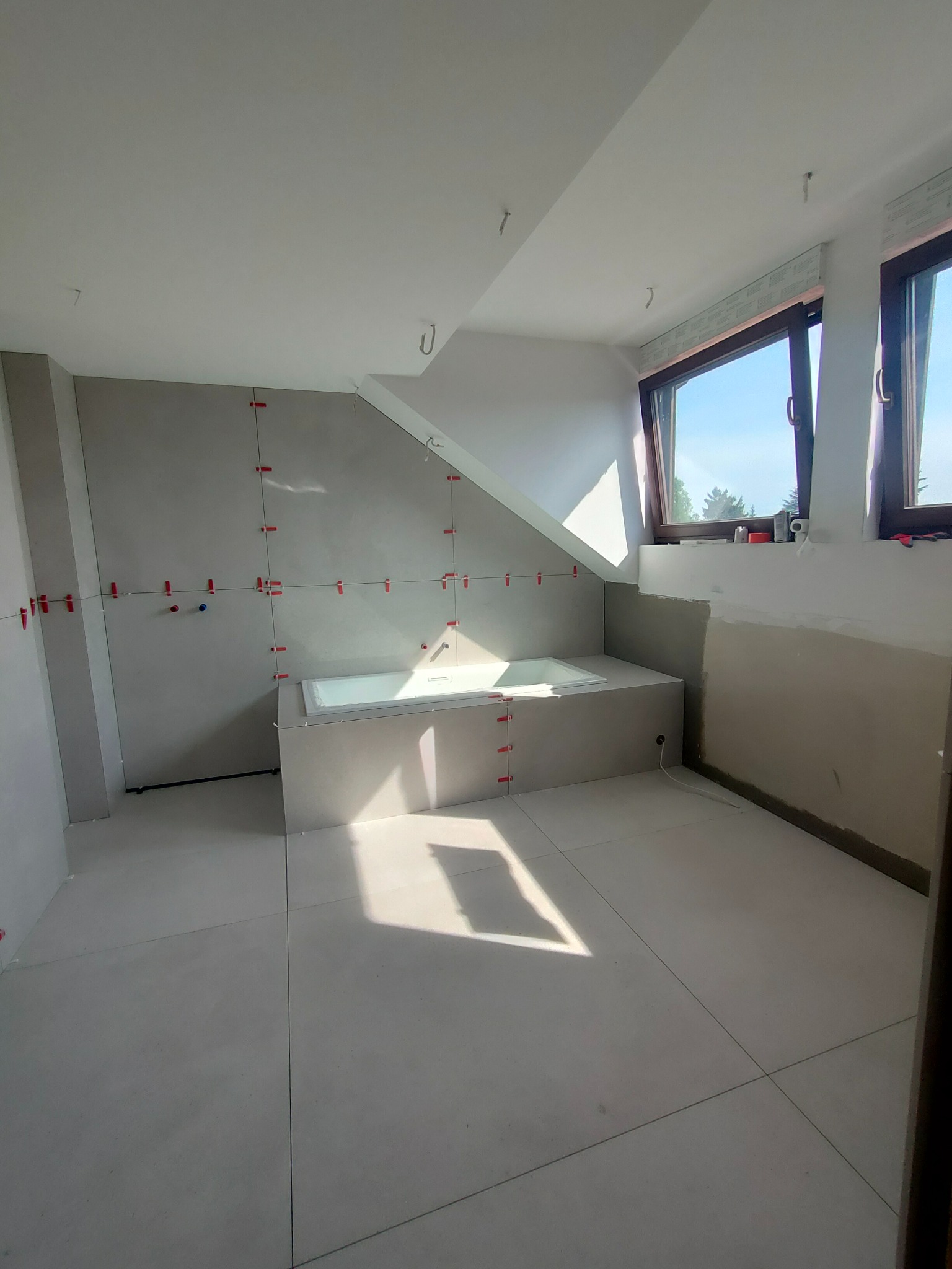 Łazienka w trakcie wykańczania: szare płytki na podłodze i ścianach, czerwone krzyżyki dystansowe, wanna obudowana płytami, okna dachowe w skosie.