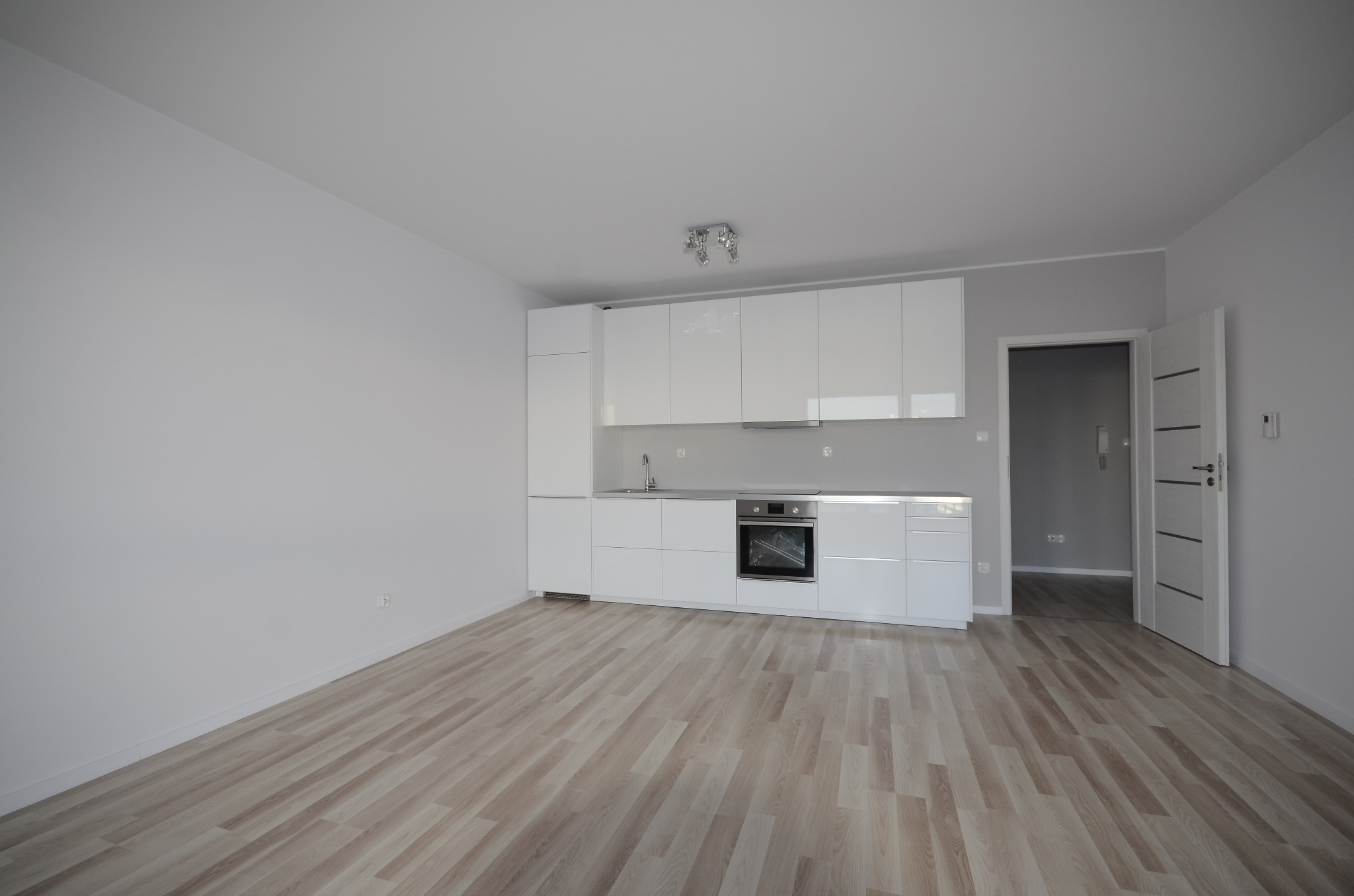 Nowoczesna, minimalistyczna kuchnia z białymi, lakierowanymi frontami szafek i drewnianą podłogą. Otwarta przestrzeń, widok na sąsiednie pomieszczenie przez otwarte drzwi.