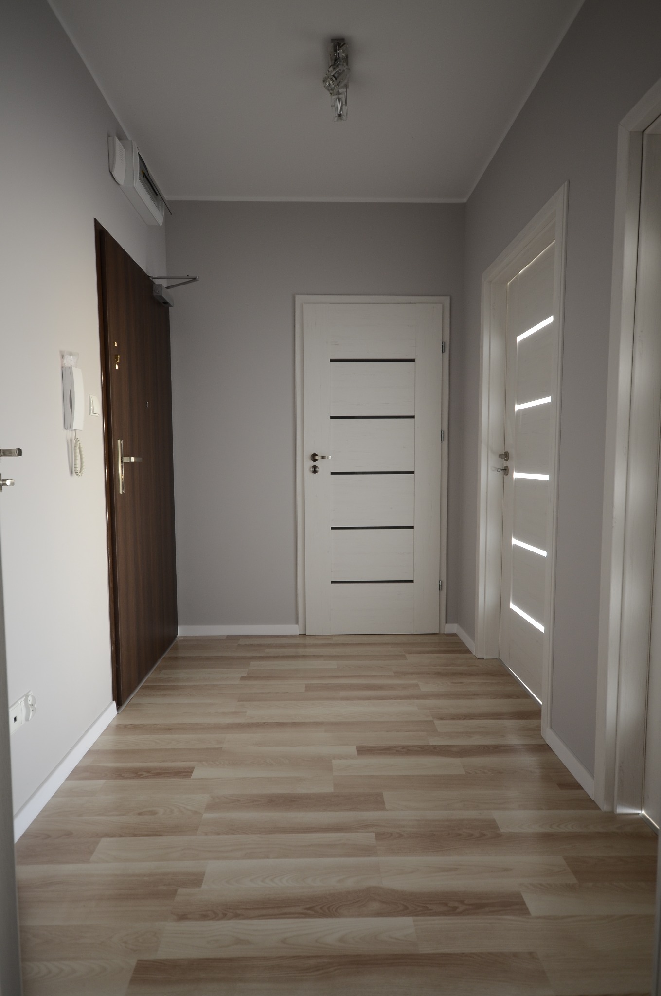 Jasne wnętrze przedpokoju z trzema parami drzwi: ciemne wejściowe, białe z poziomymi frezami i białe z pionowymi. Podłoga wyłożona panelami imitującymi drewno.