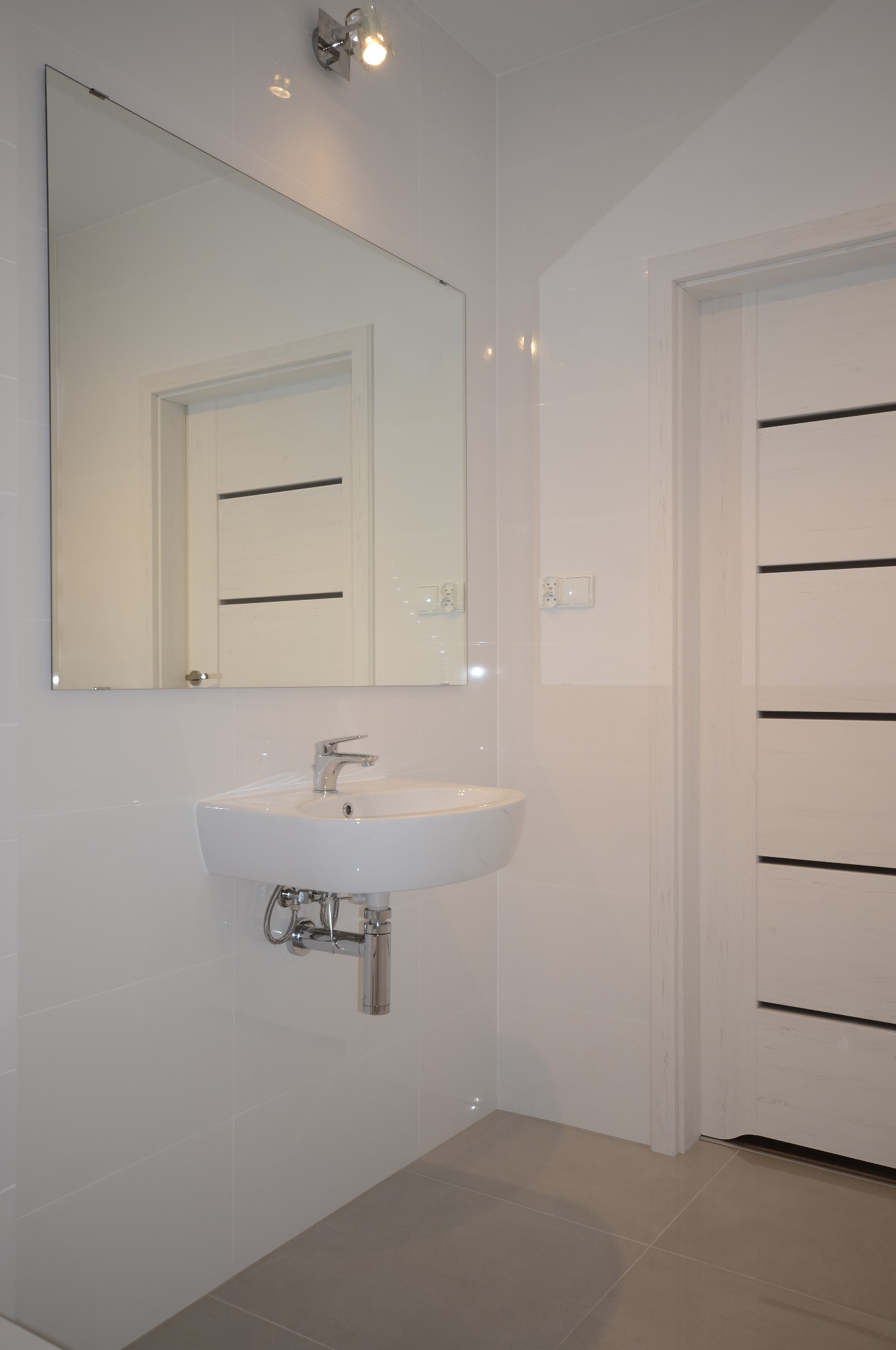 Biała, nowoczesna łazienka z wiszącą umywalką, dużym lustrem i drzwiami z poziomymi panelami. Szare płytki podłogowe i błyszczące, białe płytki ścienne.