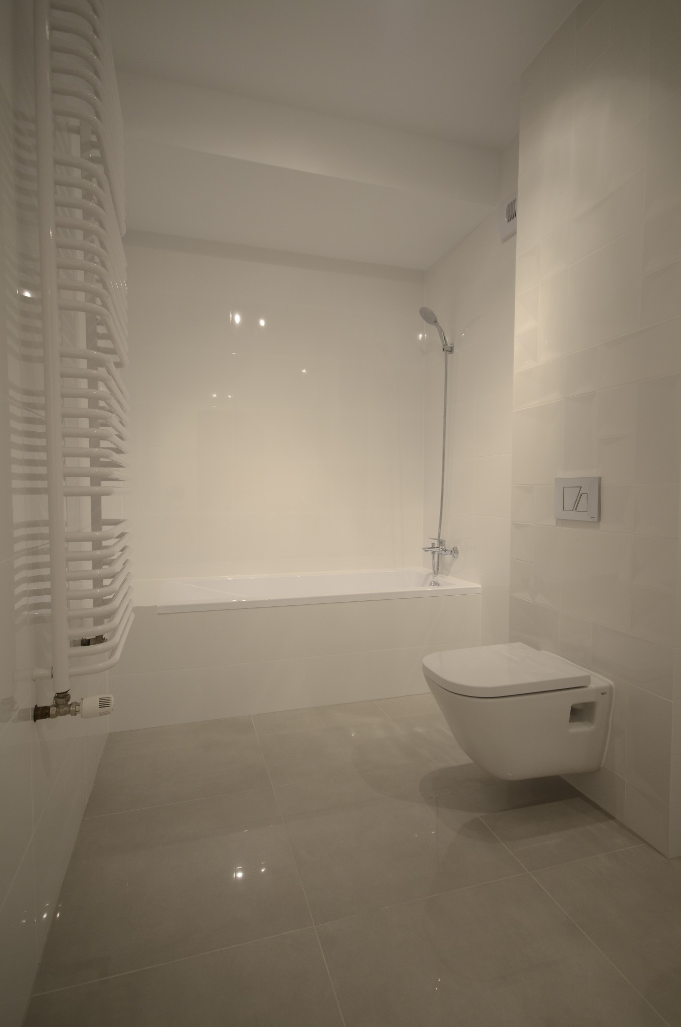 Minimalistyczna, jasna łazienka z wanną, podwieszaną toaletą i grzejnikiem drabinkowym. Błyszczące płytki podłogowe odbijają światło. Nowoczesny design.