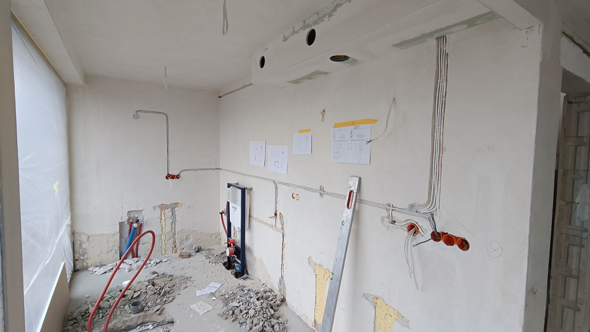 Wnętrze w trakcie remontu: widoczne instalacje elektryczne i hydrauliczne, poziomica oparta o ścianę, plany na ścianie. Fragmentarycznie usunięte stare tynki.