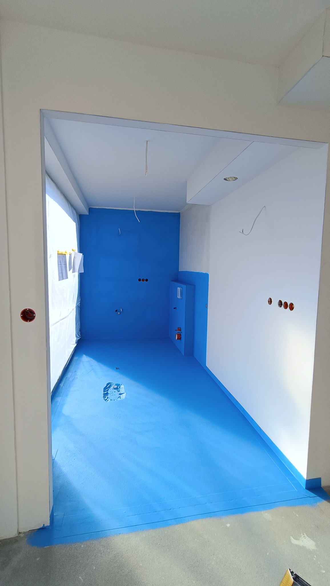 Niebieska hydroizolacja w łazience przed wykończeniem. Widoczne przygotowane ściany, podłoga i zabudowa podtynkowa. Otwory na gniazdka i instalacje.