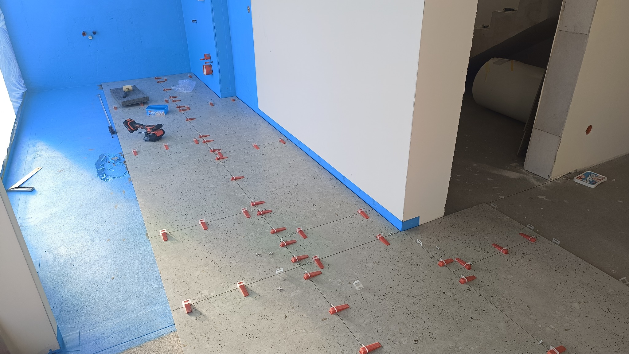 Układanie płytek podłogowych w pomieszczeniu z niebieską hydroizolacją. Widoczne narzędzia i system poziomowania płytek. Etap prac wykończeniowych.